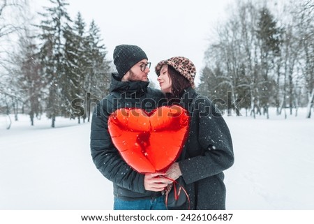 Happy beautiful young couple of lovers in fashionable winter clothes holding a red heart balloon and walking in a winter park with snow