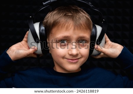 portrait of smiling fair-haired teenage boy in blue wearing headphones, looking straight ahead, holding headphones. In a dark, soundproof room
