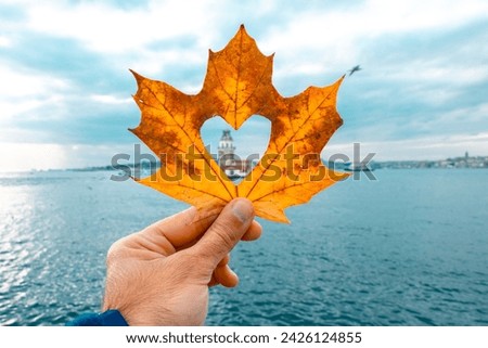 Photo of the Maiden's Tower on the Bosphorus. The man is holding the leaf in his hand. Photo of the tower with an autumn leaf cut into a heart shape. The tower is blurred in the photo.