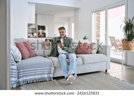 Smiling relaxed senior mature middle aged man holding cell mobile phone using smartphone sitting at home on couch in modern living room, scrolling social media, buying online, texting messages. Royalty-Free Stock Photo #2426125831