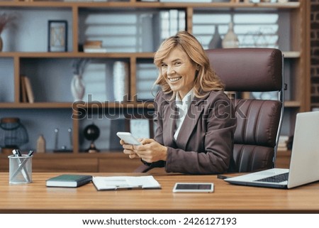 Mature businesswoman with a pleasant smile working remotely using a laptop and smartphone in a well-appointed home office.