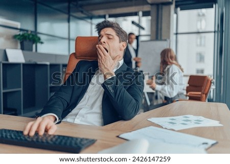 Young overtired businessman yawning while working inside modern office with computer Royalty-Free Stock Photo #2426147259