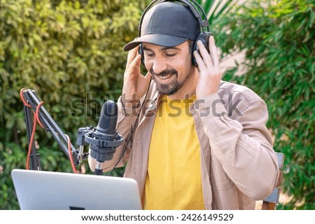 A man with a beard and moustache wearing a cap and headphones is singing and recording podcast into a microphone while using a laptop outdoor. He looks happy and relaxed, enjoying a leisurely musical