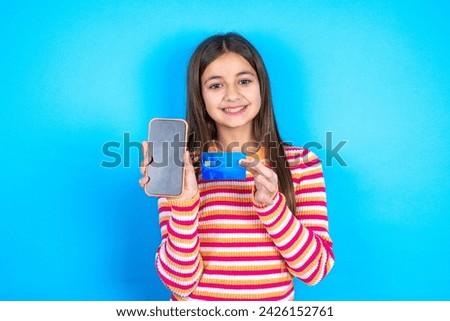 Photo of adorable beautiful kid girl wearing striped t-shirt holding credit card and Smartphone. Reserved for online purchases