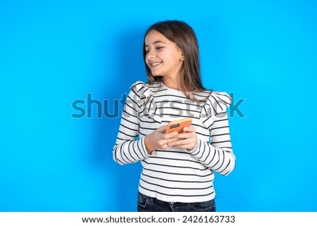 beautiful kid girl wearing striped t-shirt over blue background holding a smartphone and looking sideways at blank copyspace.