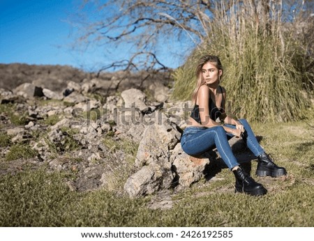 Nature photography on a summer sunny day: Young woman surrounded by nature, trees, green plants and rocks. Wearing a urban casual outfit with dark blue jeans and black crop top with black boots 