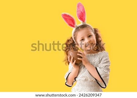 Cute little girl in bunny ears with chocolate Easter egg on yellow background Royalty-Free Stock Photo #2426209347