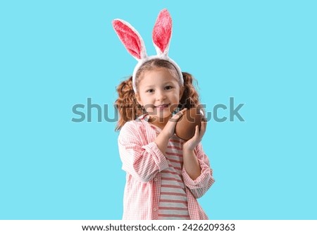 Cute little girl in bunny ears with chocolate Easter egg on blue background Royalty-Free Stock Photo #2426209363