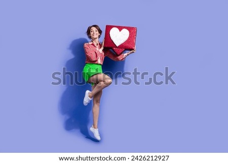 Full size photo of satisfied impressed woman dressed print shirt holding social media heart box jumping isolated on blue color background
