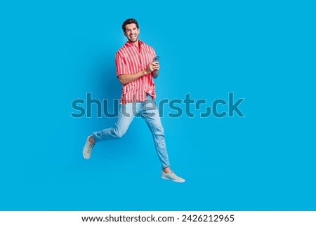 Full size photo of intelligent man dressed striped shirt denim pants run empty space hold smartphone isolated on blue color background