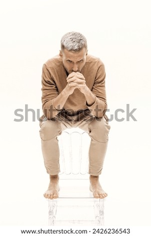 A guy in a beige jumper and cargo pants sits on a transparent chair on a white background.