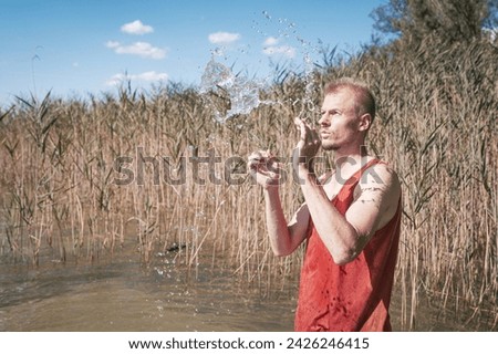 A man wearing a muscle shirt stands on the shore of a lake and has scooped water into the air. Royalty-Free Stock Photo #2426246415