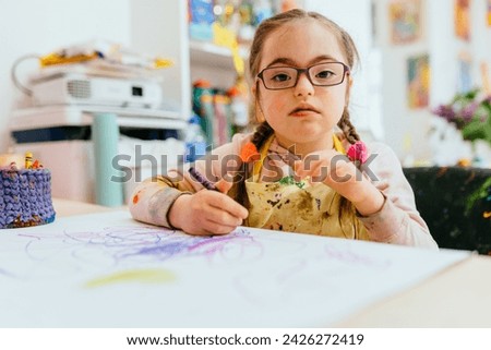 A little child girl who have down syndrome, paints a drawing in an album and smiles with a dirty face and hands, a happy child draws a picture at home at the table