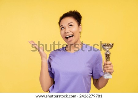 Portrait of positive beautiful African American woman, winner holding trophy cup, looking up standing isolated on yellow background. Victory concept