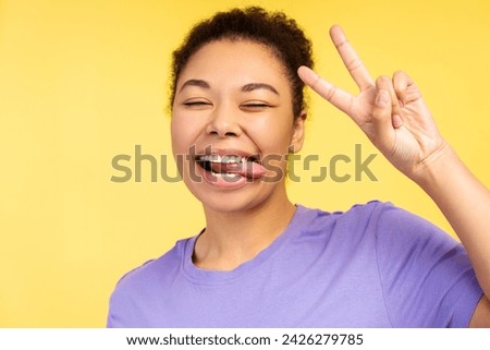 Portrait of smiling African American woman showing tongue, gesturing peace gesture, fooling around standing isolated on yellow background, closeup. Positive lifestyle concept