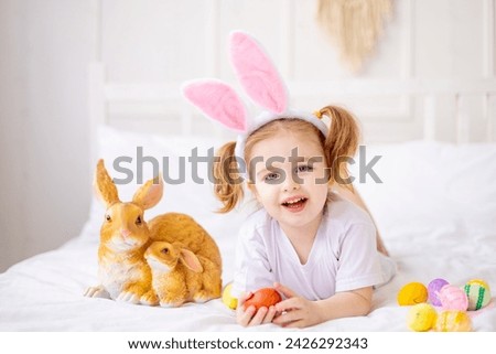 a cute little girl with bunny ears and colorful eggs on a white bed at home playing and smiling, a blonde child celebrates happy Easter