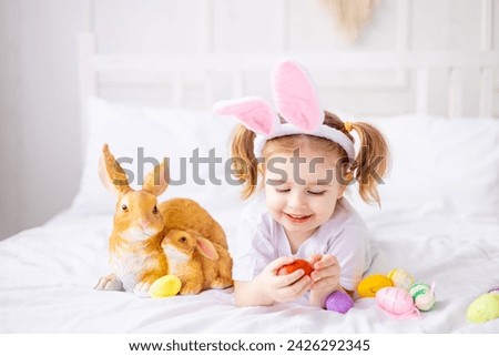 a cute little girl with bunny ears and colorful eggs on a white bed at home playing and smiling, a blonde child celebrates happy Easter