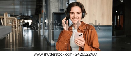 Portrait of happy young woman, manager in office, sitting in lounge room, relaxing on lunch in break room, holding smartphone, listening music in wireless headphones, using co-working space.