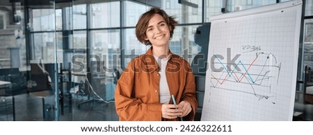 Image of young saleswoman, start up manager stands near board with diagram, giving presentation to team in office and smiling. Royalty-Free Stock Photo #2426322611