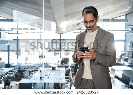 Busy mature business man ceo investor wearing suit using mobile cell phone. Older businessman executive or entrepreneur holding smartphone looking at cellphone standing in corporate office.
