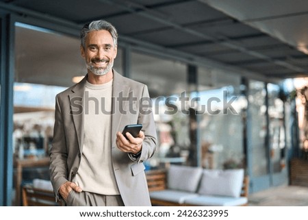 Happy mature older business man entrepreneur wearing suit using mobile cell phone outdoors. Middle aged businessman executive holding smartphone looking at camera standing outside office. Portrait. Royalty-Free Stock Photo #2426323995