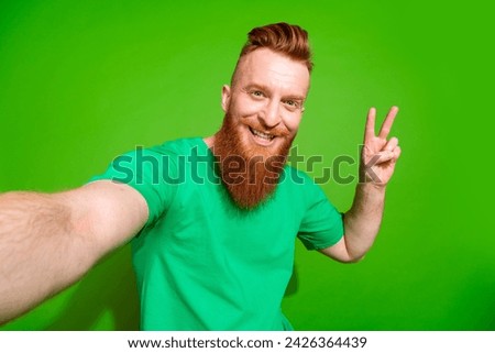 Portrait of pleasant cool guy with long red beard wear stylish t-shirt making selfie show v-sign isolated on green color background