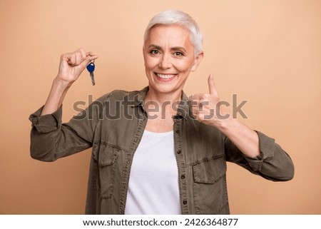 Photo portrait of pretty senior female hold key show thumb up wear trendy khaki outfit isolated on beige color background