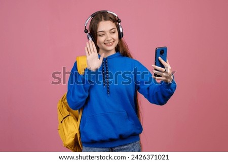 Front view of pretty brunette girl with earphones standing, taking selfie. Stylish young student with earphones, smiing, waving, greeting. Isolated on pink studio background. Royalty-Free Stock Photo #2426371021