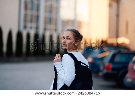 A beautiful blonde teenager returns home from school during the sunset, carrying her school backpack, in a serene and peaceful urban setting