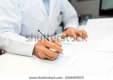 cardiologist looking at electrocardiograph ekg paper filling patient form. sitting and writing notes at the desk in clinic or hospital office Royalty-Free Stock Photo #2426405015