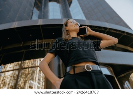 Smiling business woman in eyeglases is talking mobile phone while standing on building background