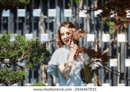 A beautiful young girl in a light summer polka dot dress stands in a park near a plant. The time of the year is summer Royalty-Free Stock Photo #2426467015