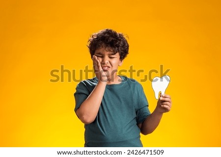 Portrait of child boy holding papercraft tooth, touching cheek because of toothache , over yellow background. Dental health concept
