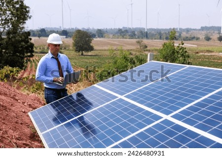 Technician specialist examining solar panel and hold using digital laptop computer in solar farm. wind turbine to generate electricity background.