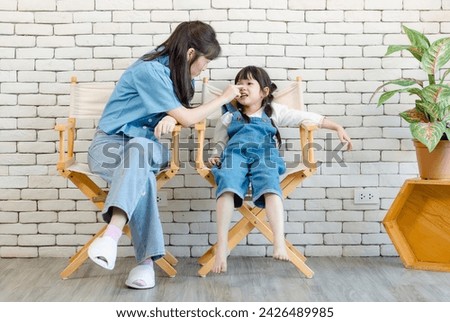 Happy asian beautiful young mother cute daughter little girl posing smiling and sitting on chair, white brick wall background studio portrait Mother's Day love family parenthood childhood concept.