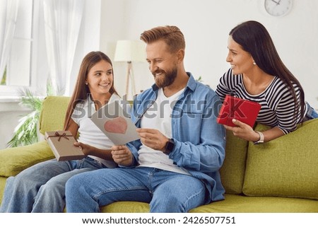 Cheerful father sitting on sofa at home with hir girl child and wife and reading handmade greeting card with heart during holiday celebration fathers day. Happy man dad receiving present from family.