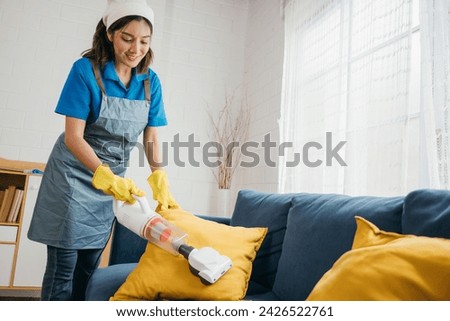 In a cozy living room an Asian woman maid meticulously vacuum machine cleaners a sofa emphasizing hygiene. Her dedication to housework and furniture care using modern technology is evident. Royalty-Free Stock Photo #2426522761