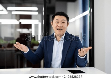 Asian businessman in a suit gesticulates while explaining a concept or giving a presentation in a modern office setting. Royalty-Free Stock Photo #2426528337