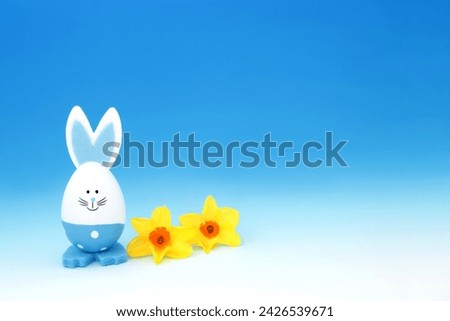 Easter bunny blue egg with Spring narcissus flowers on gradient blue white background. Minimal cute  fun composition with for the holiday season.