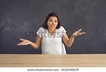 Young caucasian unsure woman sitting at a table shrugs saying that she can't help you. Beautiful brunette woman making a gesture to the camera which means I don't know. Grey studio background. Royalty-Free Stock Photo #2426540639
