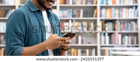Banner of smiling Indian student chatting by mobile phone in university library. Young man using social media application app, browsing online on smartphone cellphone device gadget for studying. 
