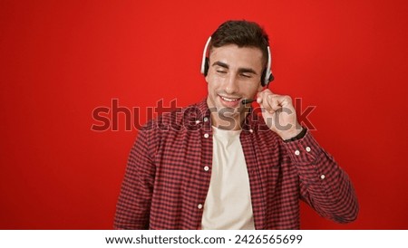 Dashing young hispanic man smiling wide, emanating confidence as a professional call center agent. wearing headset at work, over isolated red background. Royalty-Free Stock Photo #2426565699