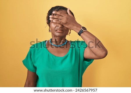 African woman with dreadlocks standing over yellow background covering eyes with hand, looking serious and sad. sightless, hiding and rejection concept  Royalty-Free Stock Photo #2426567957
