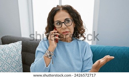 A mature hispanic woman with curly hair talking on a phone in a cozy living room, showing confusion or concern. Royalty-Free Stock Photo #2426572717