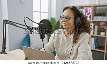 A smiling mature hispanic woman wearing headphones speaks into a studio microphone while reading from a tablet indoors.