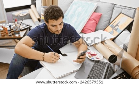 Young man sketching in an art studio while checking his smartphone, surrounded by paintings and art supplies.
