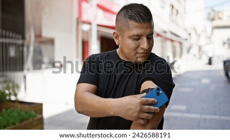 Confident young latin man, happily scanning his diabetes sensor on the city street using his smartphone, expertly monitoring his glucose levels, putting technology to work Royalty-Free Stock Photo #2426588191
