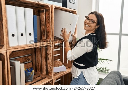 Young beautiful hispanic woman business worker holding documents of shelving at office