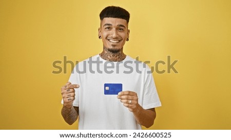 Confident young latin man, smiling cheerfully, pointing at his credit card, isolated on a vibrant yellow background. positive financial expression of wealth and joy.