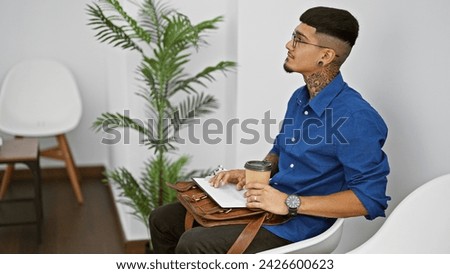Handsome young latin man with a tattoo, seriously sitting in the waiting room holding a cup of well-deserved coffee and business briefcase, anxiously awaiting his professional interview Royalty-Free Stock Photo #2426600623